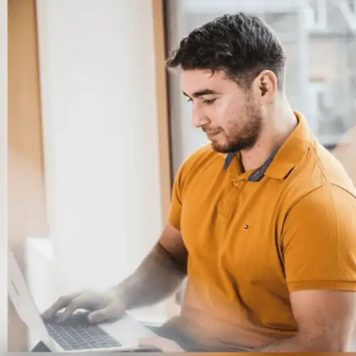 Ein Mann mit kurzen Haaren und Bart, der ein senfgelbes Poloshirt trägt, sitzt an einem Fenster und benutzt einen Laptop. Er wirkt konzentriert und beschäftigt sich mit dem Webdesign und der Entwicklung auf dem Bildschirm. Der Hintergrund ist sanft beleuchtet, was darauf hindeutet, dass natürliches Licht durch das Fenster fällt. - digiak webdesign und entwicklung